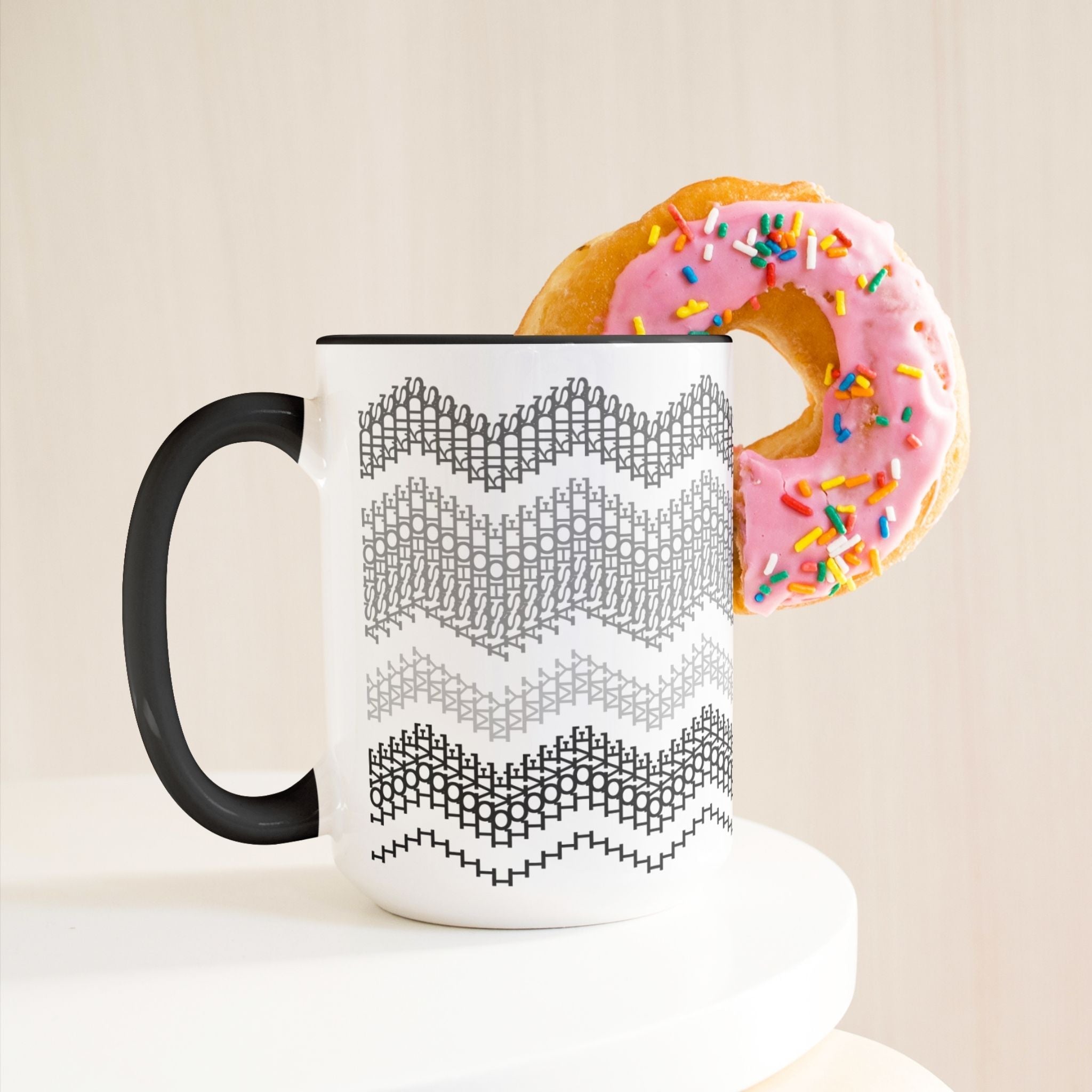 Gray &amp; Black hidden message mug 15oz on white pedestal with a pink frosted sprinkle donut leaning against it — lifestyle product shot on warm cream background