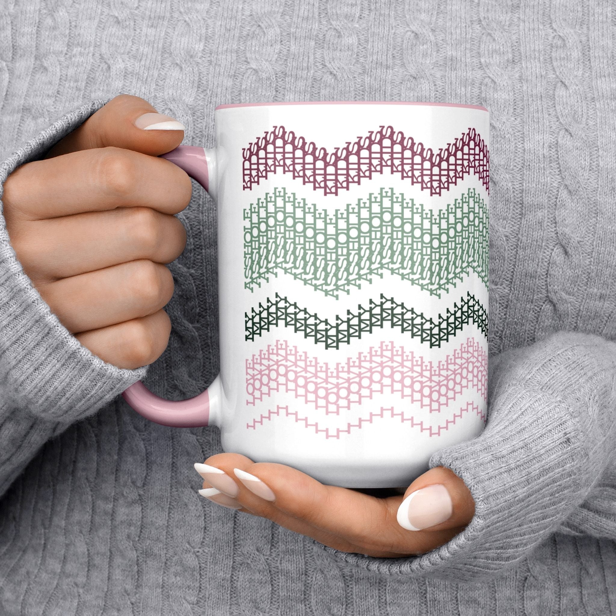Preppy &amp; Pink hidden message mug 15oz cradled in two hands wearing a gray cable-knit sweater — cozy lifestyle shot showing full typographic cross-stitch pattern in botanical tones
