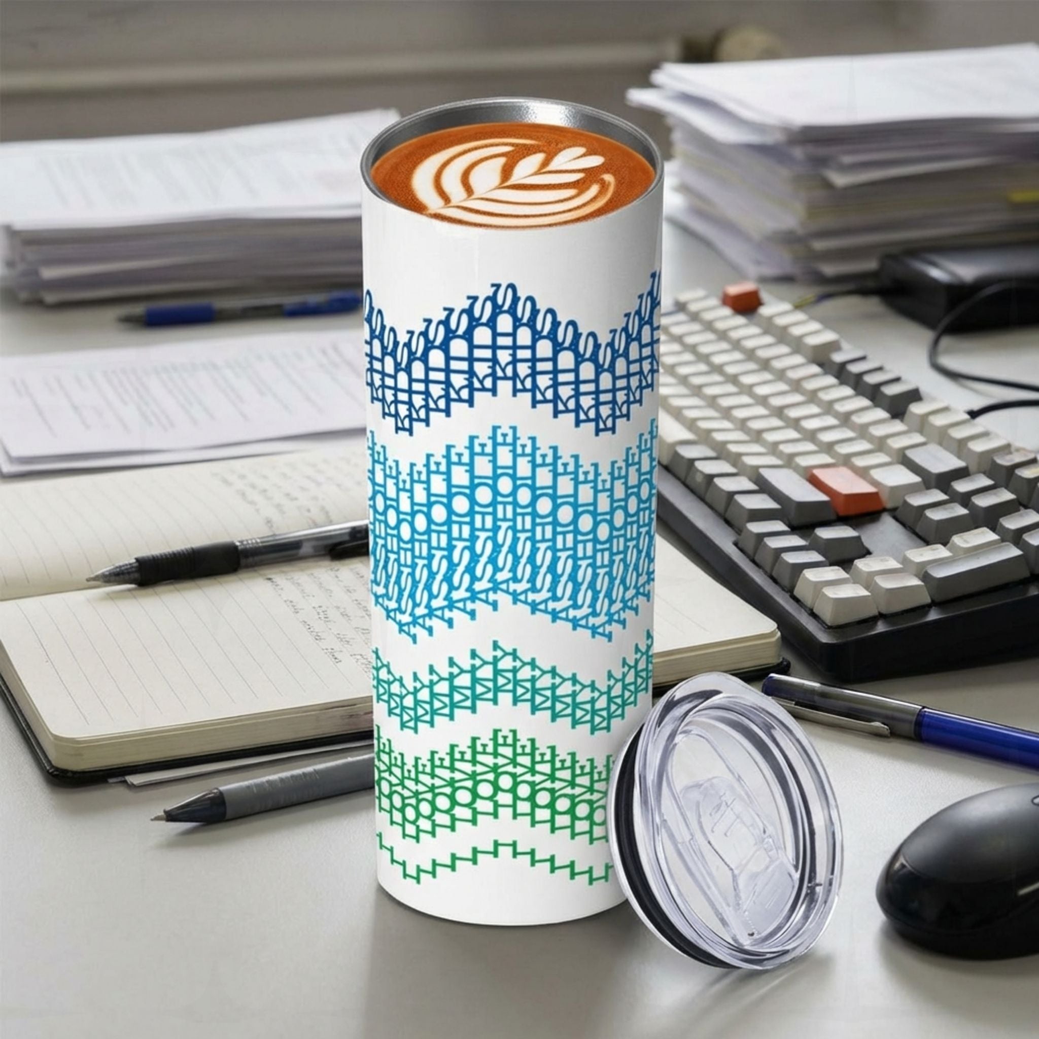 Hidden message stainless steel tumbler displayed on a work desk next to coffee equipment, ocean stripes colorway — navy, sky blue, teal, and green embroidery-style stripe bands, lid shown separately beside the tumbler