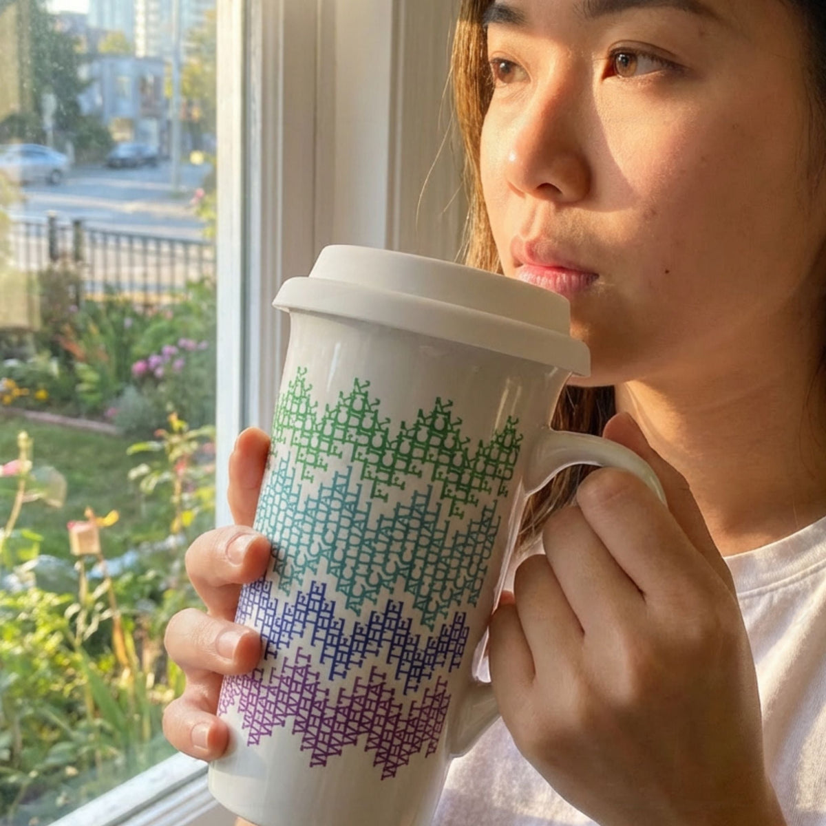 Person holding bright stripes hidden profanity latte mug by window, morning light, green teal blue purple cross-stitch text pattern visible on 16oz ceramic