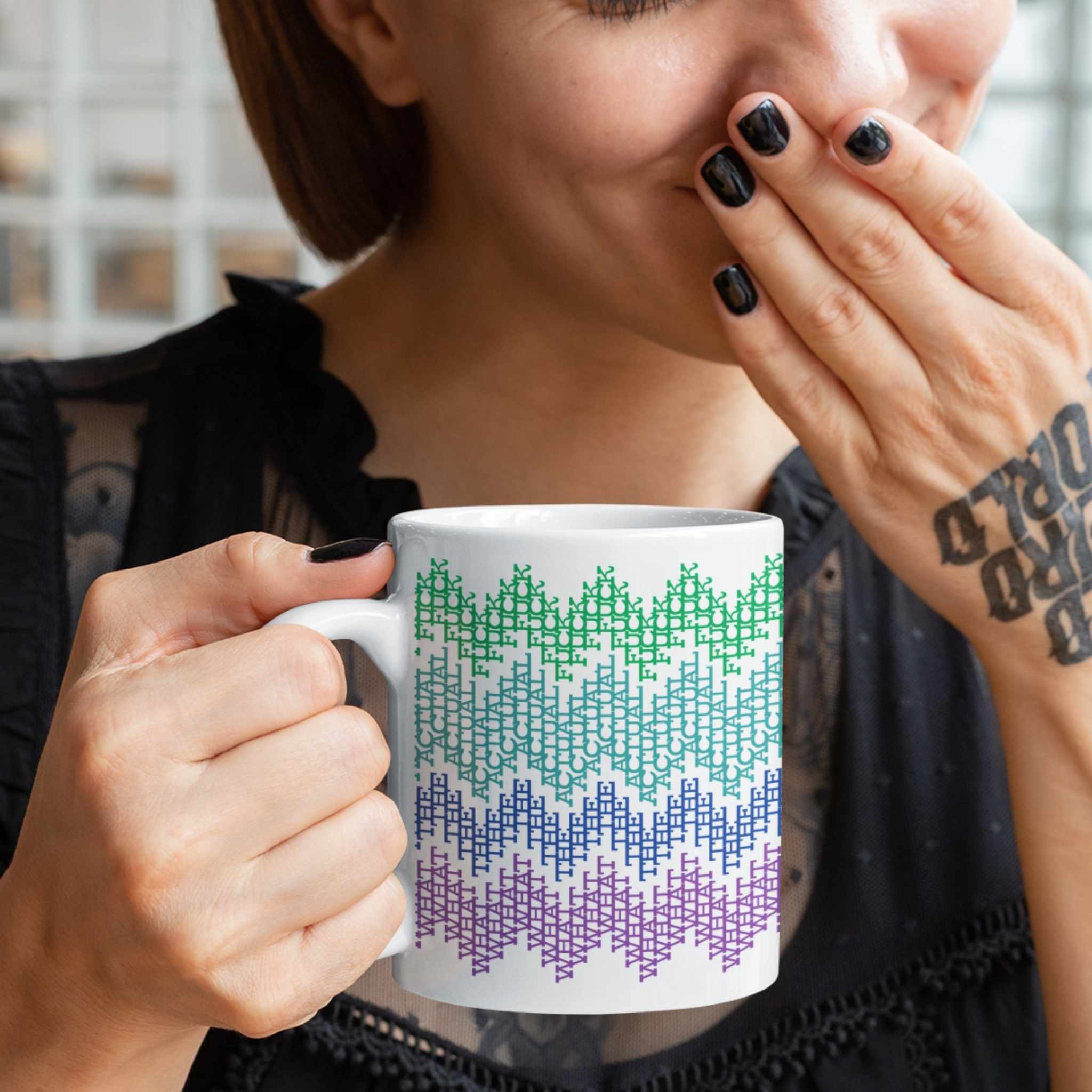 Woman with black nail polish and tattoos holding a colorful hidden profanity coffee mug while covering her mouth laughing