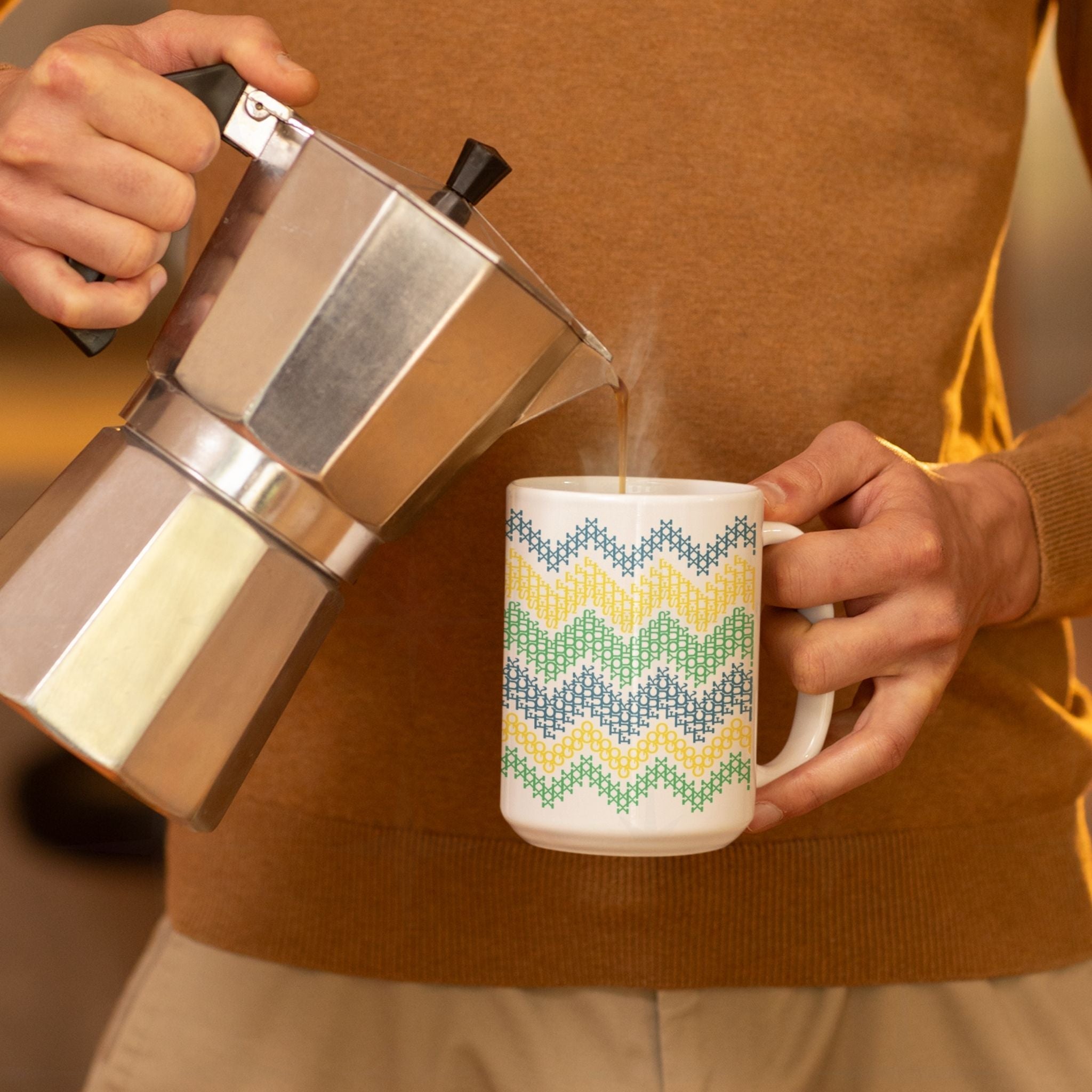 Hands pouring steaming coffee from moka pot into hidden profanity mug with bright chevron cross stitch pattern spelling "Go Fuck Your Self"