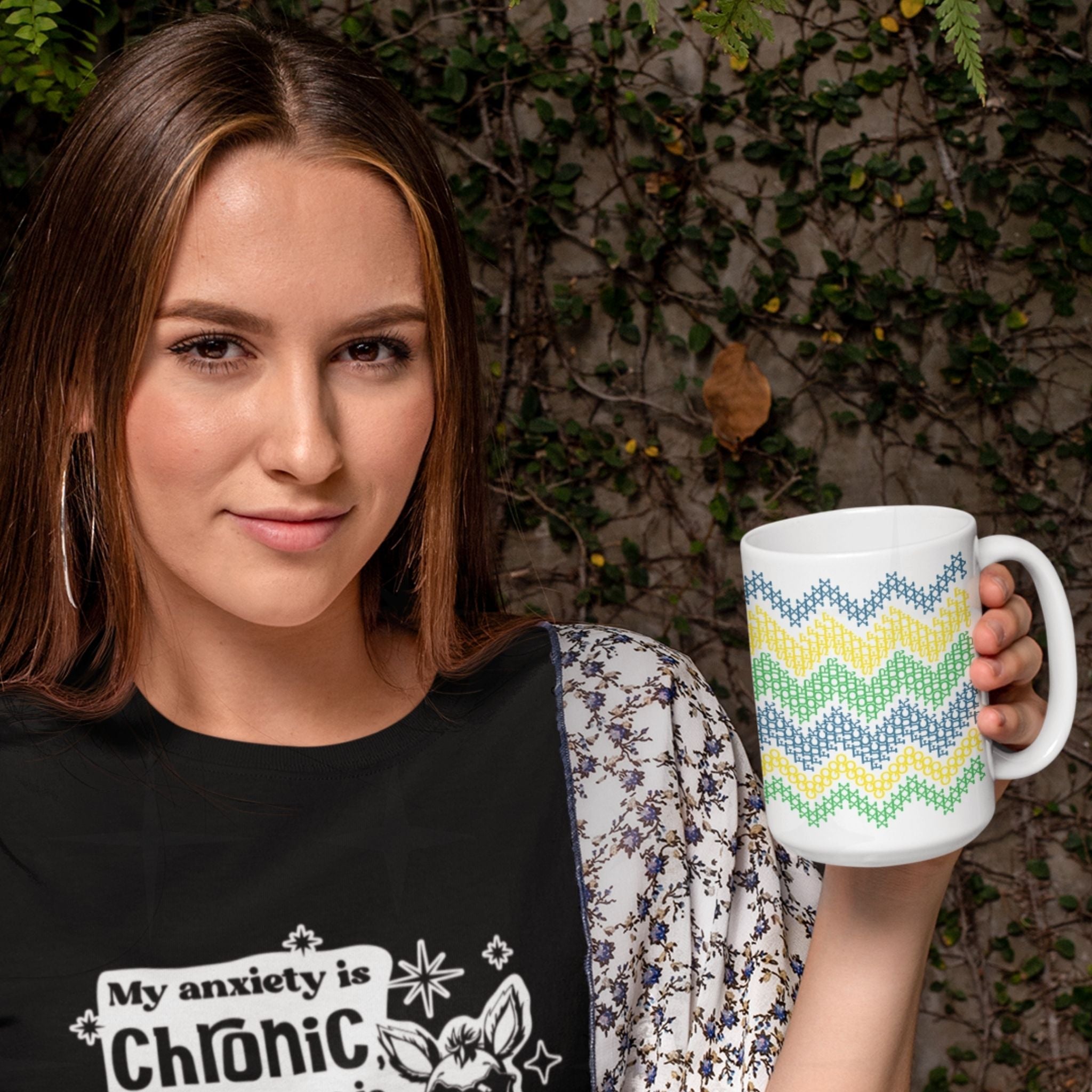 Woman holding hidden message cross stitch coffee mug with bright teal yellow green stripes outdoors against ivy wall
