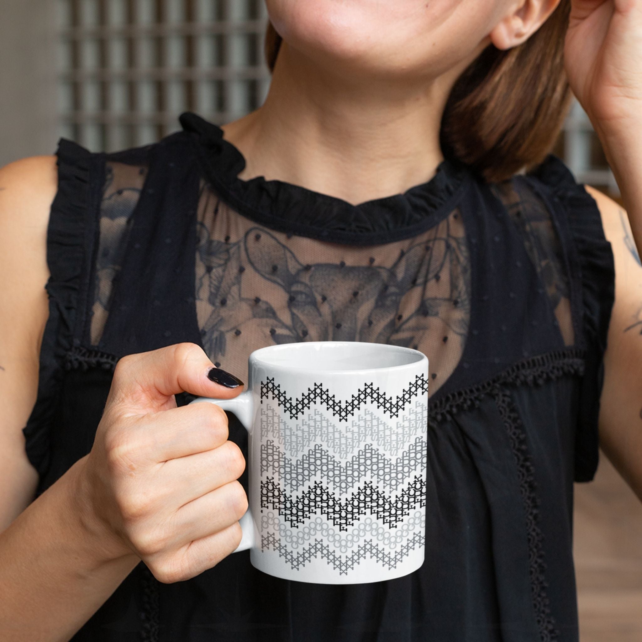 Tattooed woman in black top holding hidden profanity ceramic mug in gray cross stitch chevron pattern with black nail polish