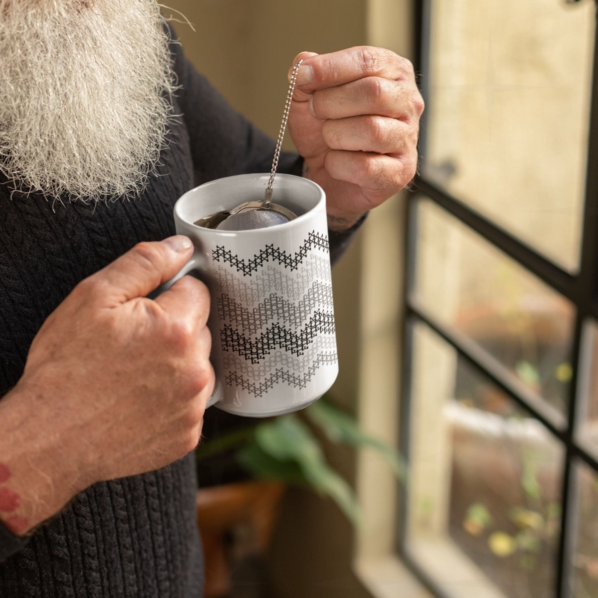 Older bearded man steeping loose leaf tea in hidden profanity ceramic mug with gray cross stitch chevron stripe pattern by window