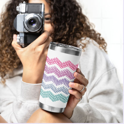 Woman with curly hair holding bright stripe hidden profanity tumbler and vintage camera showing pink purple teal cross stitch pattern