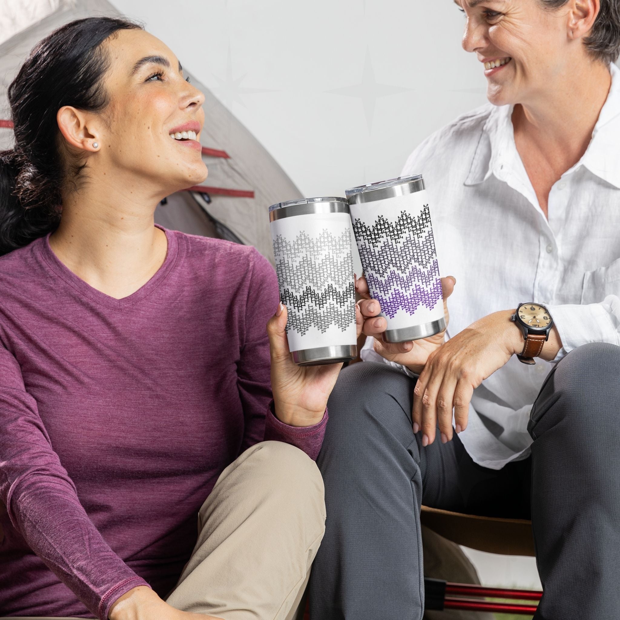 Two people sitting by a tent holding gray stripes and purple ombre hidden message tumblers, laughing together outdoors

