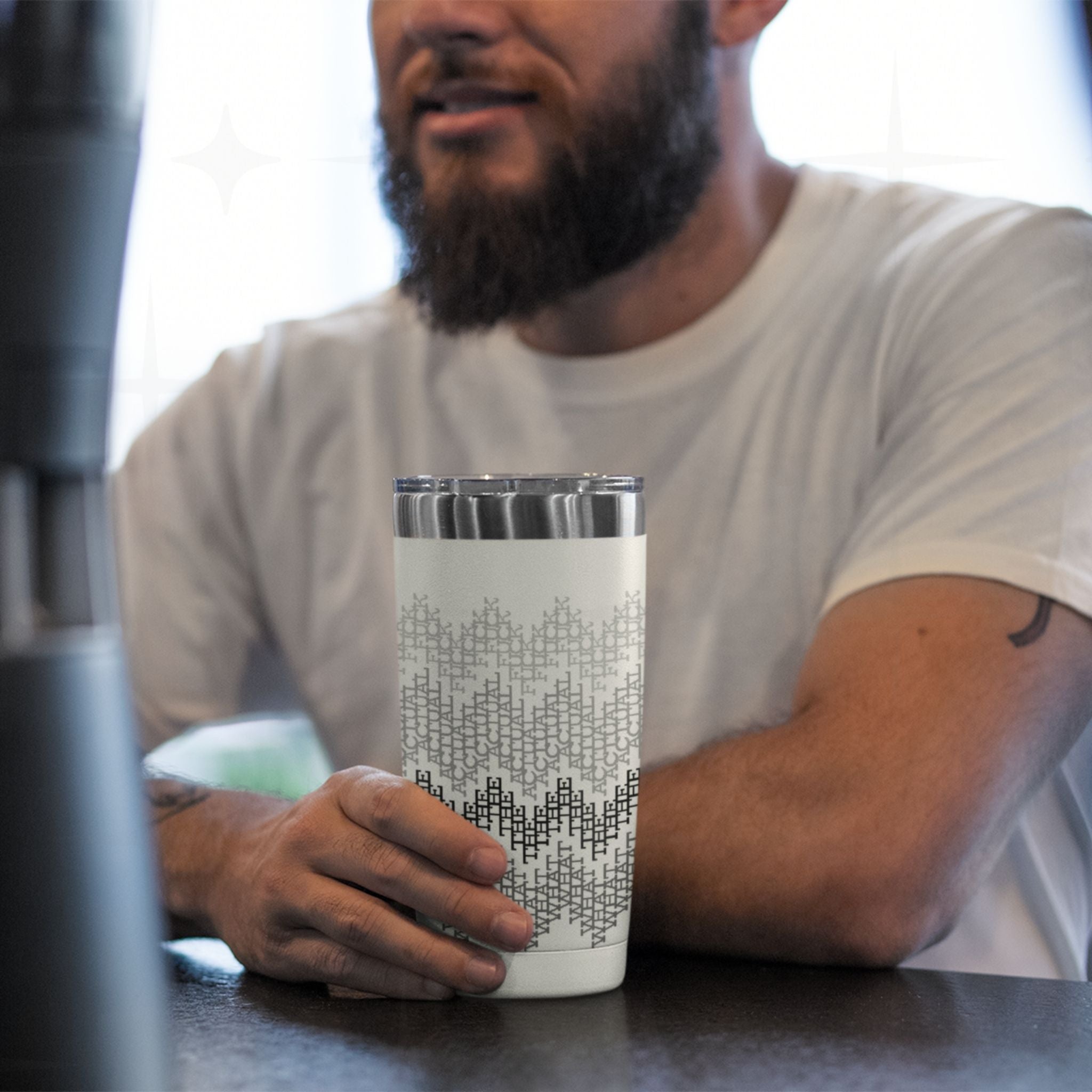 Man at kitchen counter holding gray stripes hidden message tumbler, cross-stitch swear word pattern visible on stainless steel cup