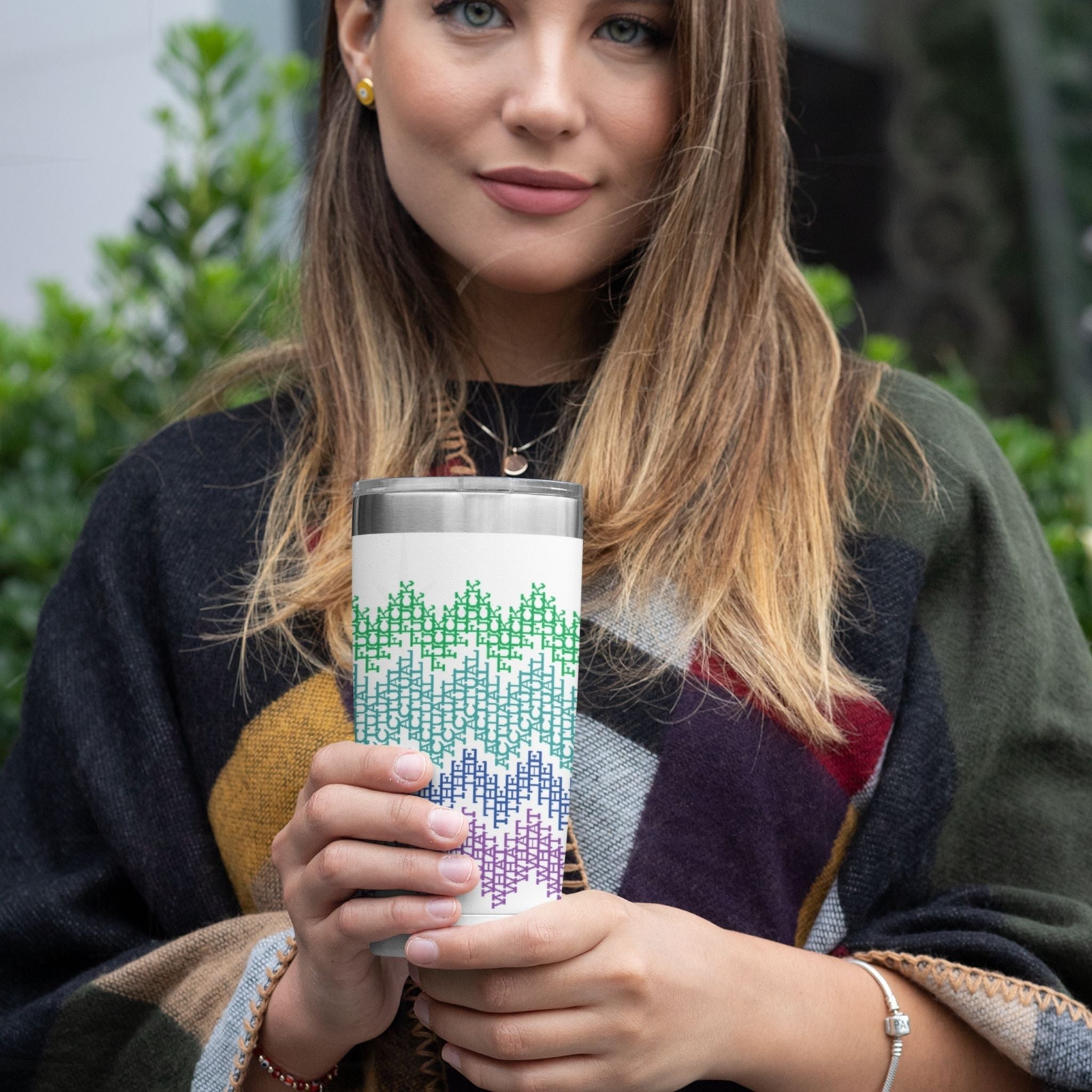 Woman holding bright stripes hidden profanity tumbler in garden setting, green teal blue purple cross-stitch pattern visible on 20oz stainless steel