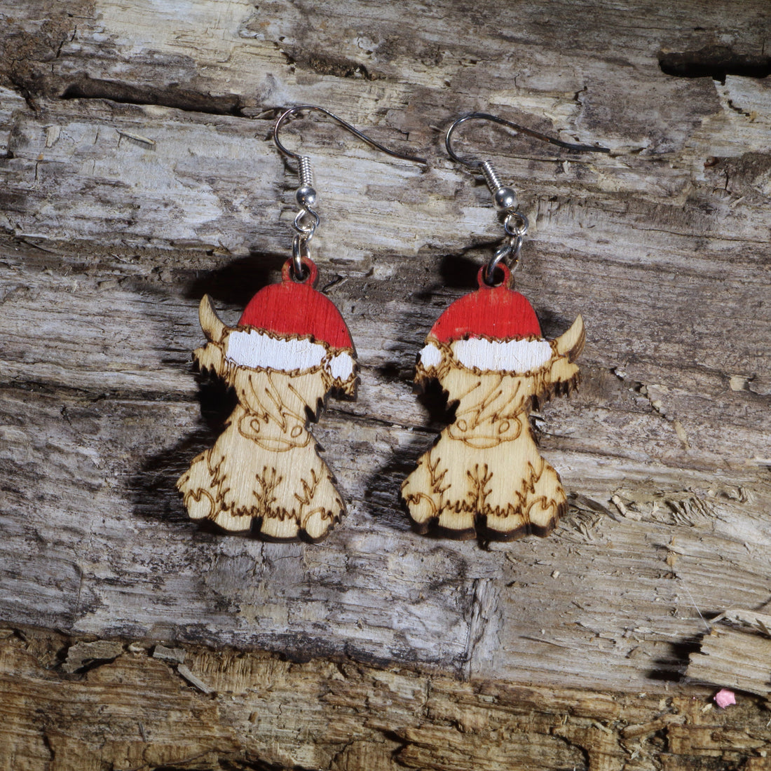 Hand-painted wooden Highland cow dangle earrings featuring an adorable fluffy Highland calf wearing a festive red and white Santa hat. Crafted from lightweight wood with hypoallergenic .925 sterling silver hooks, these charming Christmas earrings are perfect for Highland cow lovers, farm animal enthusiasts, holiday parties, winter celebrations, stocking stuffers, and anyone who loves rustic Christmas jewelry with cozy country charm.