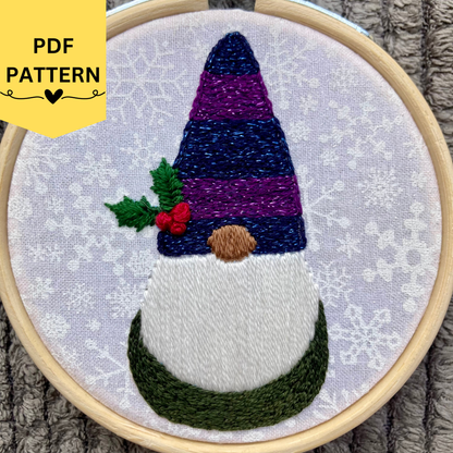 PDF pattern created by Kate, not AI. Hand embroidered Christmas gnome with a sparkle blue and purple striped winter hat and holly leaves and berries. Stitched on white cotton with sparkly white snowflakes printed on the fabric. Displayed in light wooden hoop as a DIY handmade Christmas ornament.