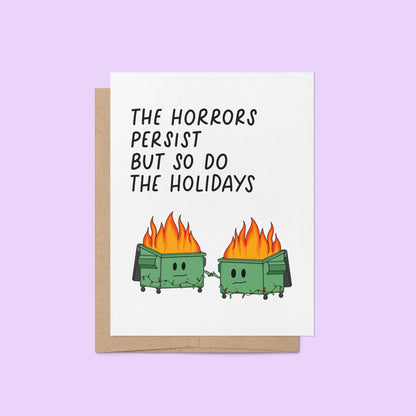 Humorous holiday card with two flaming dumpster characters holding hands, decorated with string lights, and the text “The horrors persist but so do the holidays.”