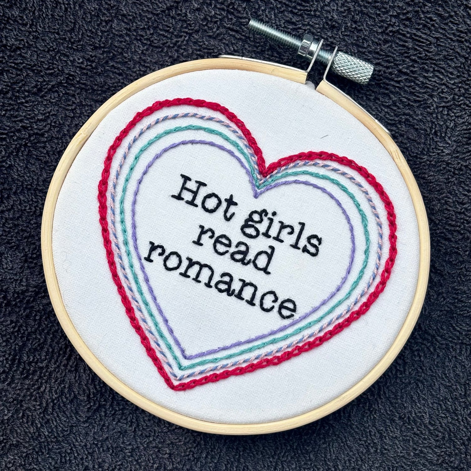 "Hot people read books" Quote hand embroidered in black embroidery thread in a serif, typewriter font. Text is framed in a series of hearts nested inside of each other and sewn in red, blue, pink, green, and purple. Each heart is stitched in a different hand embroidery stitch, as a sampler. Fabric is white cotton. Displayed as wall art in a light wooden embroidery hoop. Also sized for a christmas ornament.