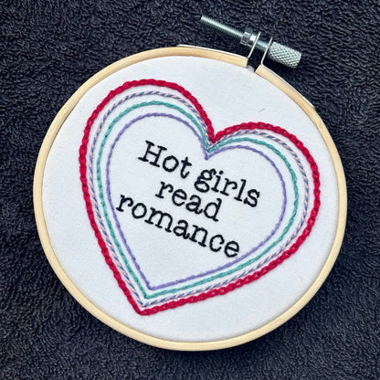 "Hot people read books" Quote hand embroidered in black embroidery thread in a serif, typewriter font. Text is framed in a series of hearts nested inside of each other and sewn in red, blue, pink, green, and purple. Each heart is stitched in a different hand embroidery stitch, as a sampler. Fabric is white cotton. Displayed as wall art in a light wooden embroidery hoop. Also sized for a christmas ornament.