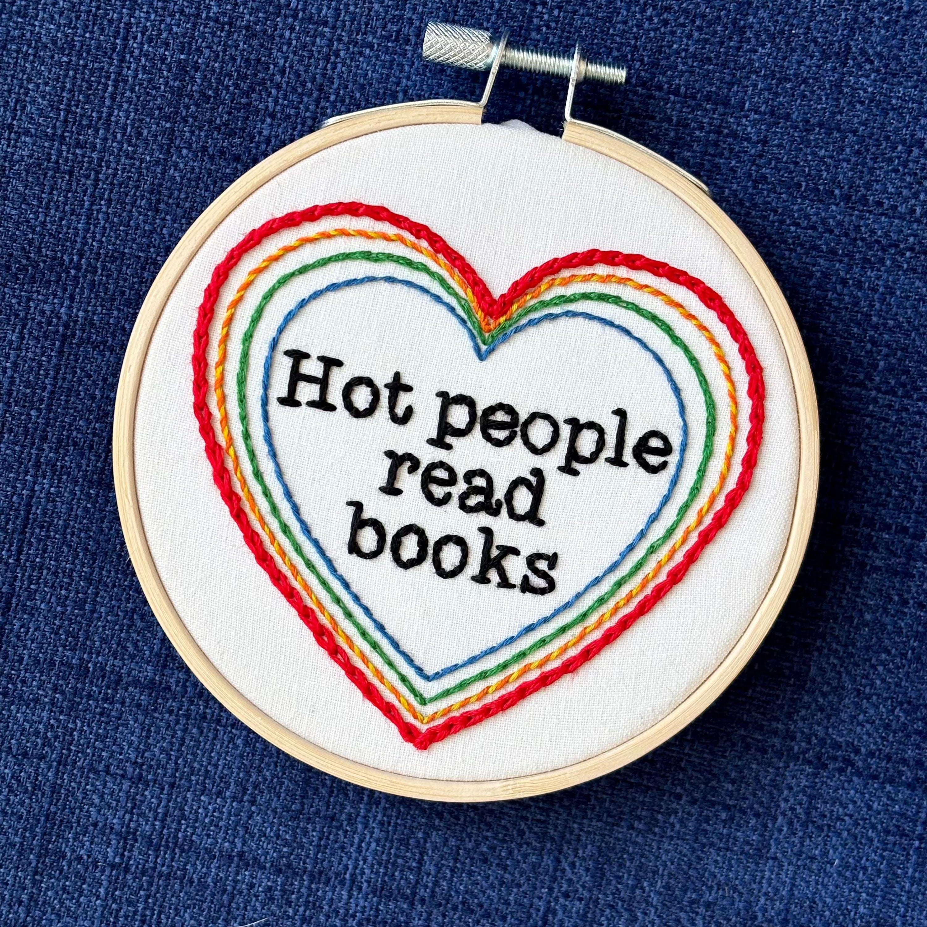 "Hot people read books" Quote hand embroidered in black embroidery thread in a serif, typewriter font. Text is framed in a series of hearts nested inside of each other and sewn in rainbow colors. Each heart is stitched in a different hand embroidery stitch, as a sampler. Fabric is white cotton. Displayed as wall art in a light wooden embroidery hoop. Also sized for a christmas ornament.