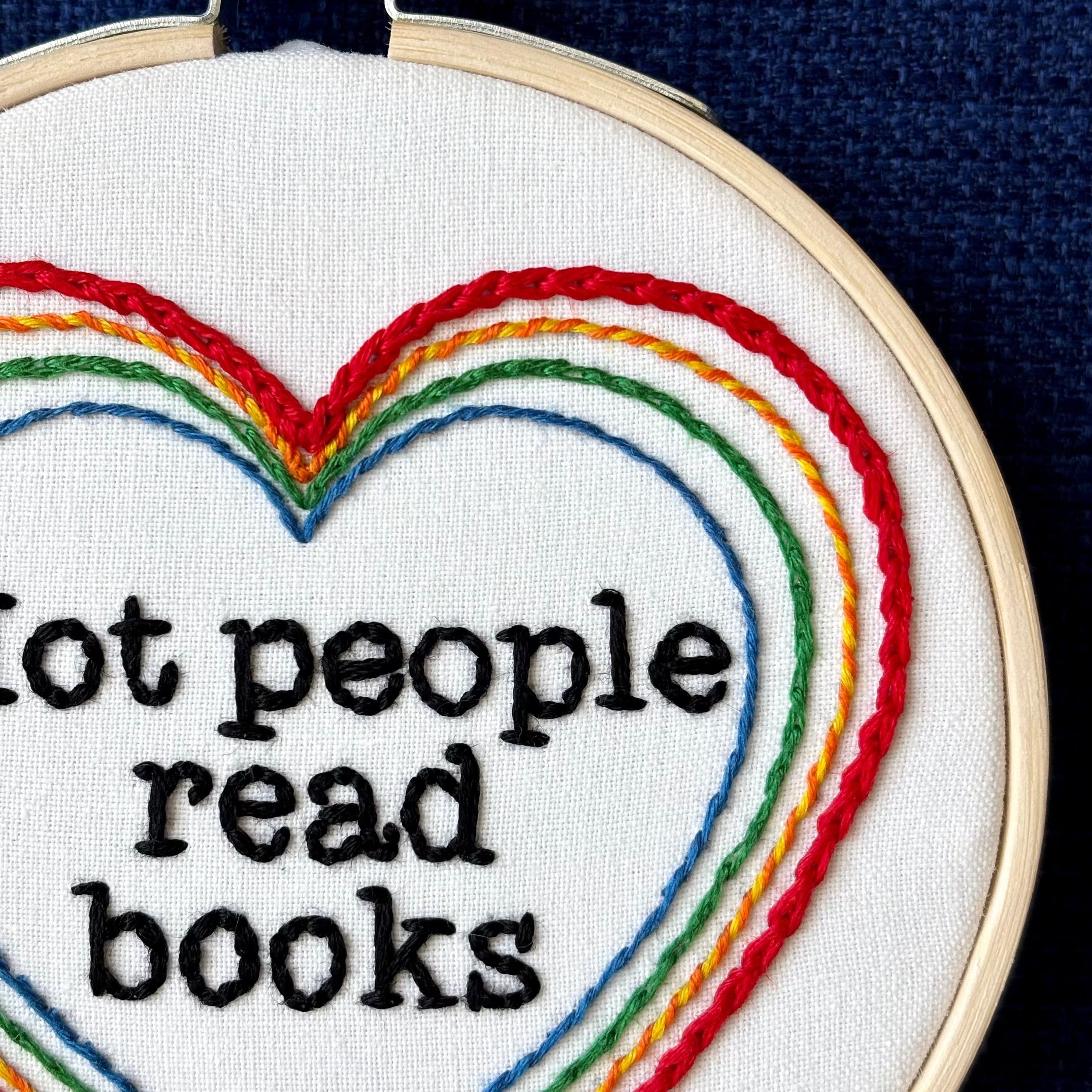 "Hot people read books" Quote hand embroidered in black embroidery thread in a serif, typewriter font. Text is framed in a series of hearts nested inside of each other and sewn in rainbow colors. Each heart is stitched in a different hand embroidery stitch, as a sampler. Fabric is white cotton. Displayed as wall art in a light wooden embroidery hoop. Also sized for a christmas ornament.