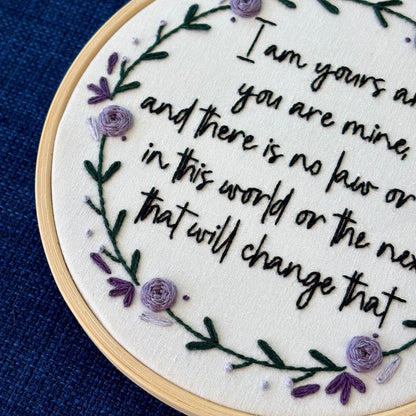 "I am yours and you are mine, and there is no law or rule in this world or the next that will change that." Quote hand embroidered in strong handwriting font in black embroidery floss. Surrounding the text is a wreath of dark green stems and leaves with lavender, violet, and deep purple flower petals, roses, and rose buds. Sewn on white cotton. Framed as wall art in a light wood embroidery hoop.