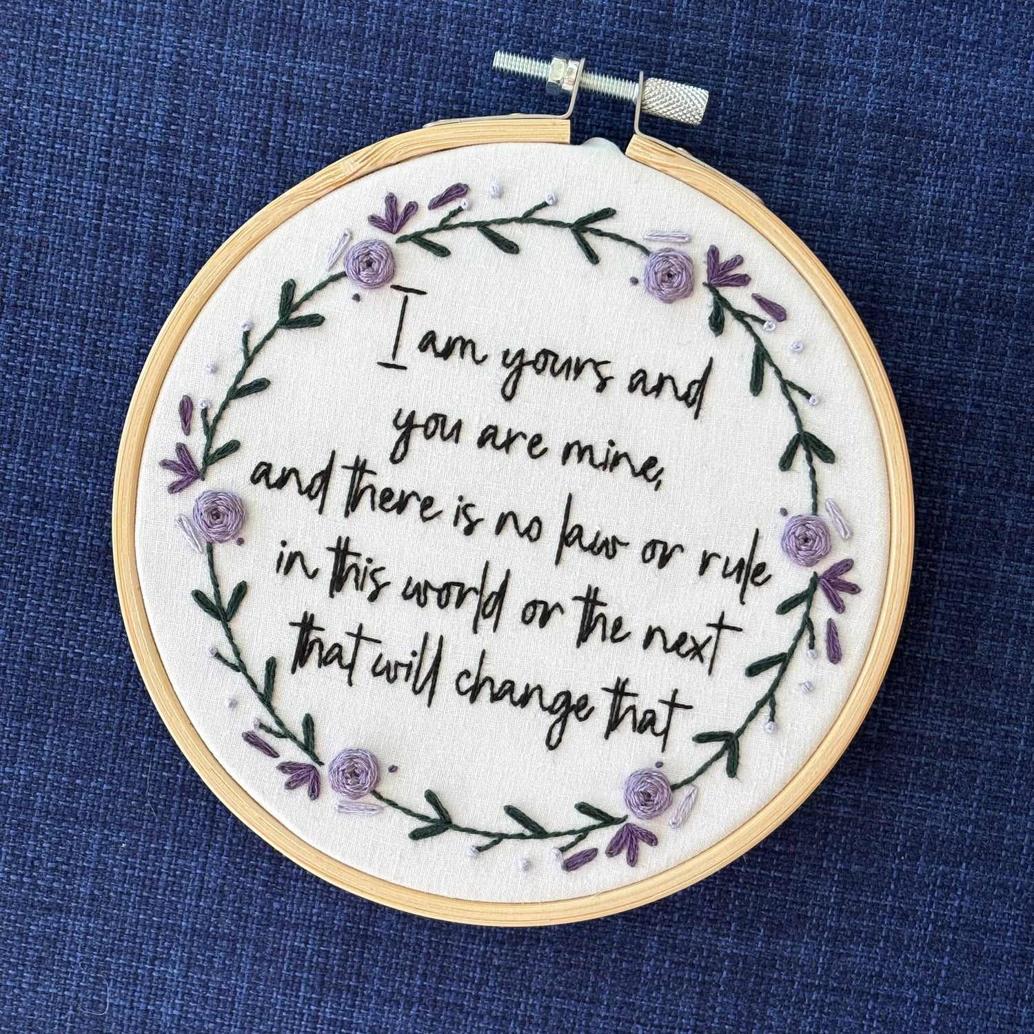 "I am yours and you are mine, and there is no law or rule in this world or the next that will change that." Quote hand embroidered in strong handwriting font in black embroidery floss. Surrounding the text is a wreath of dark green stems and leaves with lavender, violet, and deep purple flower petals, roses, and rose buds. Sewn on white cotton. Framed as wall art in a light wood embroidery hoop.