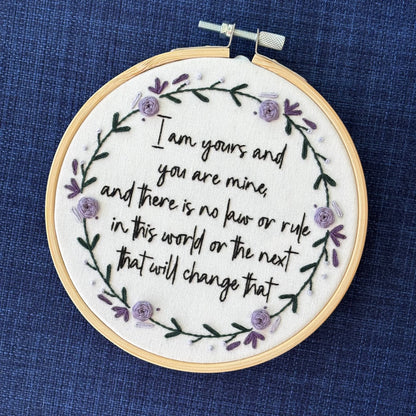 "I am yours and you are mine, and there is no law or rule in this world or the next that will change that." Quote hand embroidered in strong handwriting font in black embroidery floss. Surrounding the text is a wreath of dark green stems and leaves with lavender, violet, and deep purple flower petals, roses, and rose buds. Sewn on white cotton. Framed as wall art in a light wood embroidery hoop.