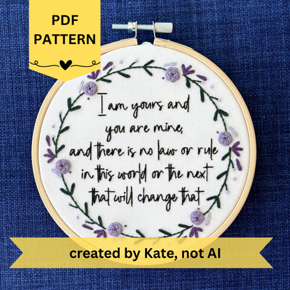 PDF pattern created by Kate, not AI. "I am yours and you are mine, and there is no law or rule in this world or the next that will change that." Quote hand embroidered in strong handwriting font in black embroidery floss. Surrounding the text is a wreath of dark green stems and leaves with lavender, violet, and deep purple flower petals, roses, and rose buds. Sewn on white cotton. Framed as wall art in a light wood embroidery hoop.