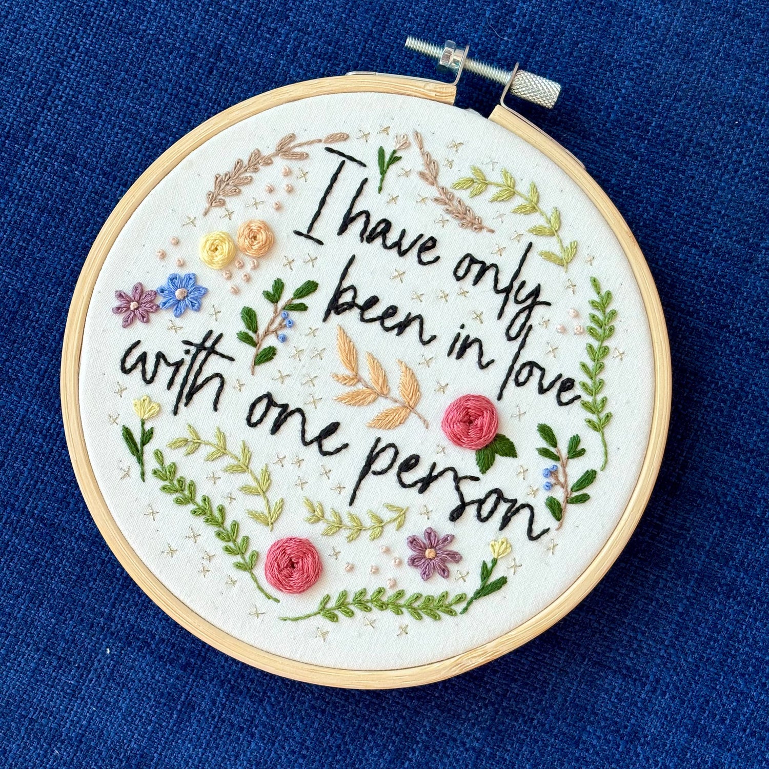 "I have only been in love with one person." Quote hand embroidered in handwriting font in black embroidery floss. Surrounding the text are vines and flowers and plants in shades of green, light browns, peach, pink, periwinkle blue, and dusky purple. Background is filled with pink dots and metallic gold Xs. Sewn on white cotton. Framed as wall art in a light wood embroidery hoop.