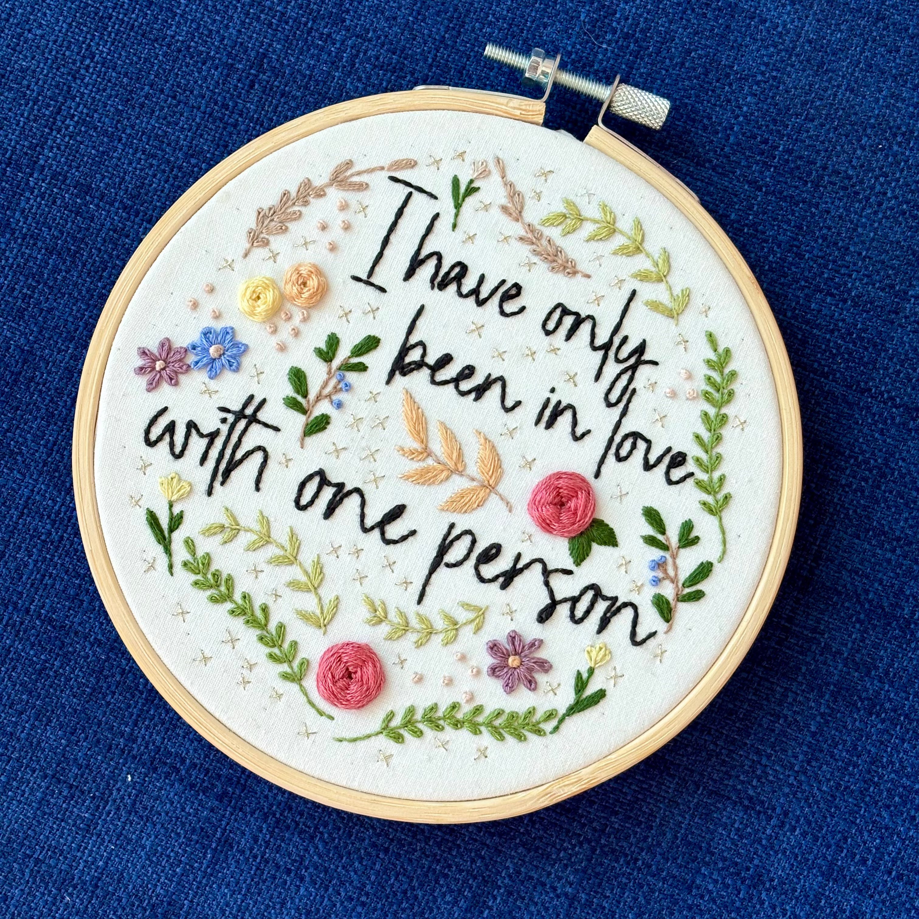 "I have only been in love with one person." Quote hand embroidered in handwriting font in black embroidery floss. Surrounding the text are vines and flowers and plants in shades of green, light browns, peach, pink, periwinkle blue, and dusky purple. Background is filled with pink dots and metallic gold Xs. Sewn on white cotton. Framed as wall art in a light wood embroidery hoop.