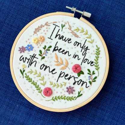"I have only been in love with one person." Quote hand embroidered in handwriting font in black embroidery floss. Surrounding the text are vines and flowers and plants in shades of green, light browns, peach, pink, periwinkle blue, and dusky purple. Background is filled with pink dots and metallic gold Xs. Sewn on white cotton. Framed as wall art in a light wood embroidery hoop.