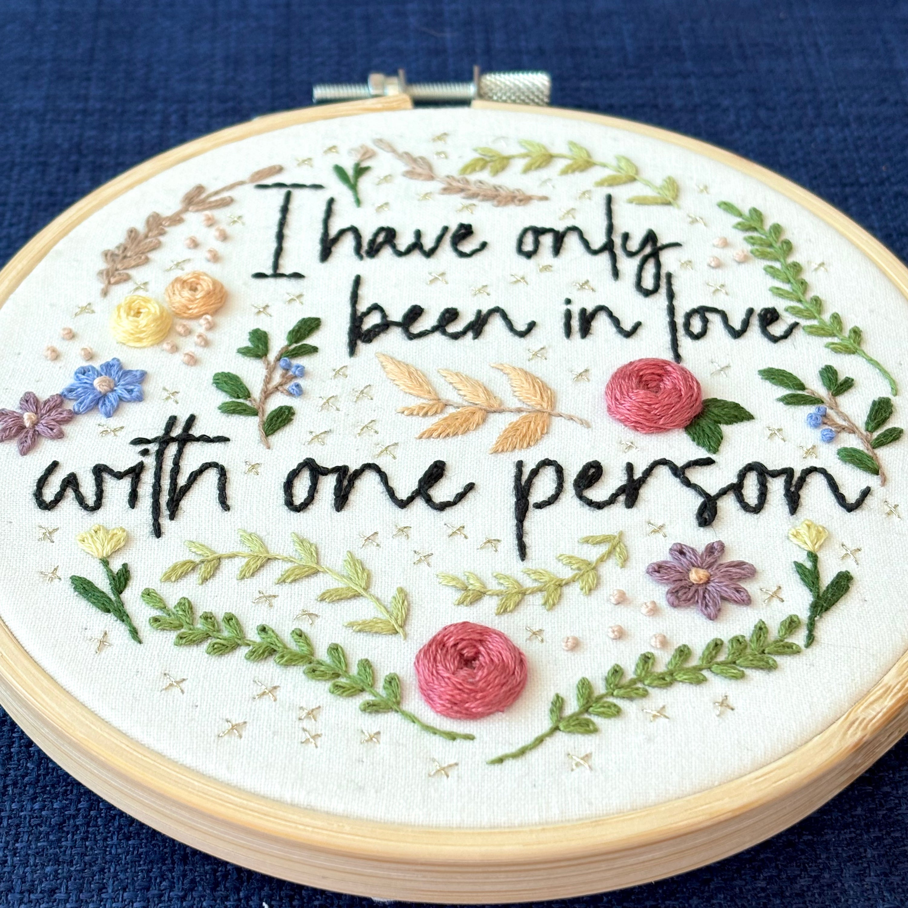 "I have only been in love with one person." Quote hand embroidered in handwriting font in black embroidery floss. Surrounding the text are vines and flowers and plants in shades of green, light browns, peach, pink, periwinkle blue, and dusky purple. Background is filled with pink dots and metallic gold Xs. Sewn on white cotton. Framed as wall art in a light wood embroidery hoop.