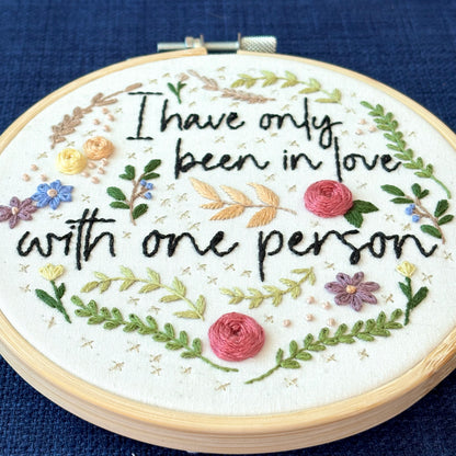 "I have only been in love with one person." Quote hand embroidered in handwriting font in black embroidery floss. Surrounding the text are vines and flowers and plants in shades of green, light browns, peach, pink, periwinkle blue, and dusky purple. Background is filled with pink dots and metallic gold Xs. Sewn on white cotton. Framed as wall art in a light wood embroidery hoop.