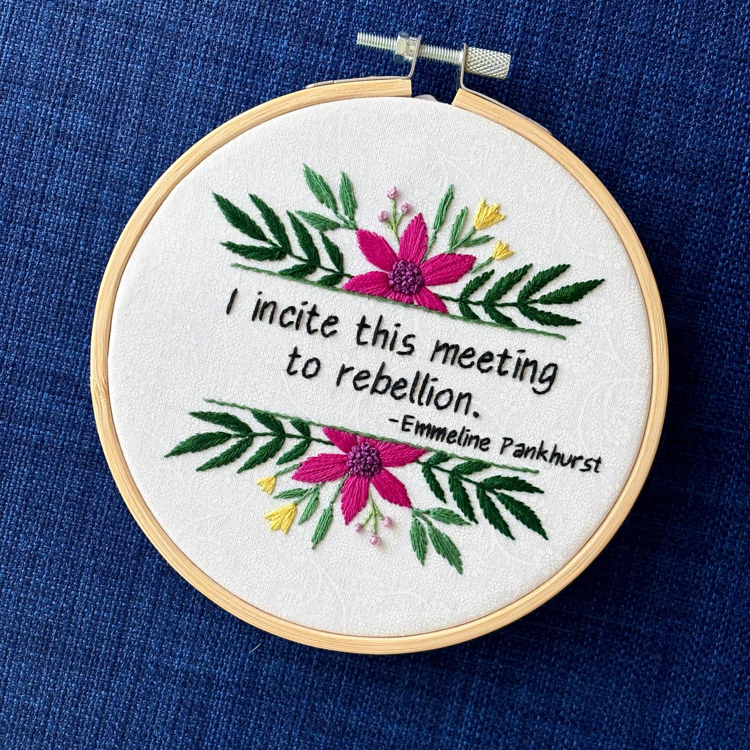 "I incite this meeting to rebellion - Emmeline Pankhurst". Quote hand embroidered in black thread. Framed with a fuchsia daisies, small yellow tulips, sprigs of green leaves, and branches with lavender flower buds. Hand sewn on white-on-white cotton and displayed as wall art in a light wooden embroidery hoop.