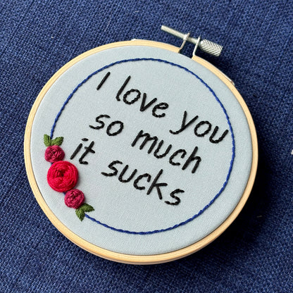 "I love you so much it sucks." Quote hand embroidered in handwriting font in black embroidery thread. Around the text is a floral wreath of a blue ring, red roses and green leaves. Fabric is light blue, and completed embroidery is displayed in a light wood embroidery hoop.