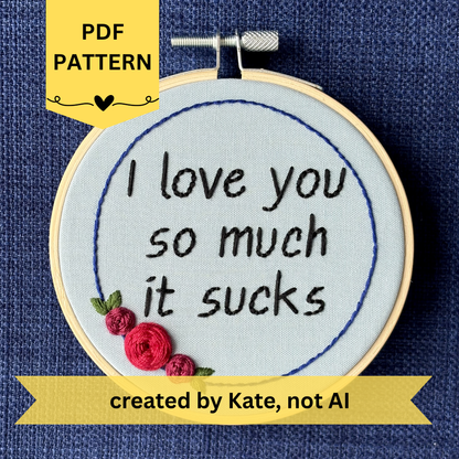 PDF pattern created by Kate, not AI. "I love you so much it sucks." Quote hand embroidered in handwriting font in black embroidery thread. Around the text is a floral wreath of a blue ring, red roses and green leaves. Fabric is light blue, and completed embroidery is displayed in a light wood embroidery hoop.