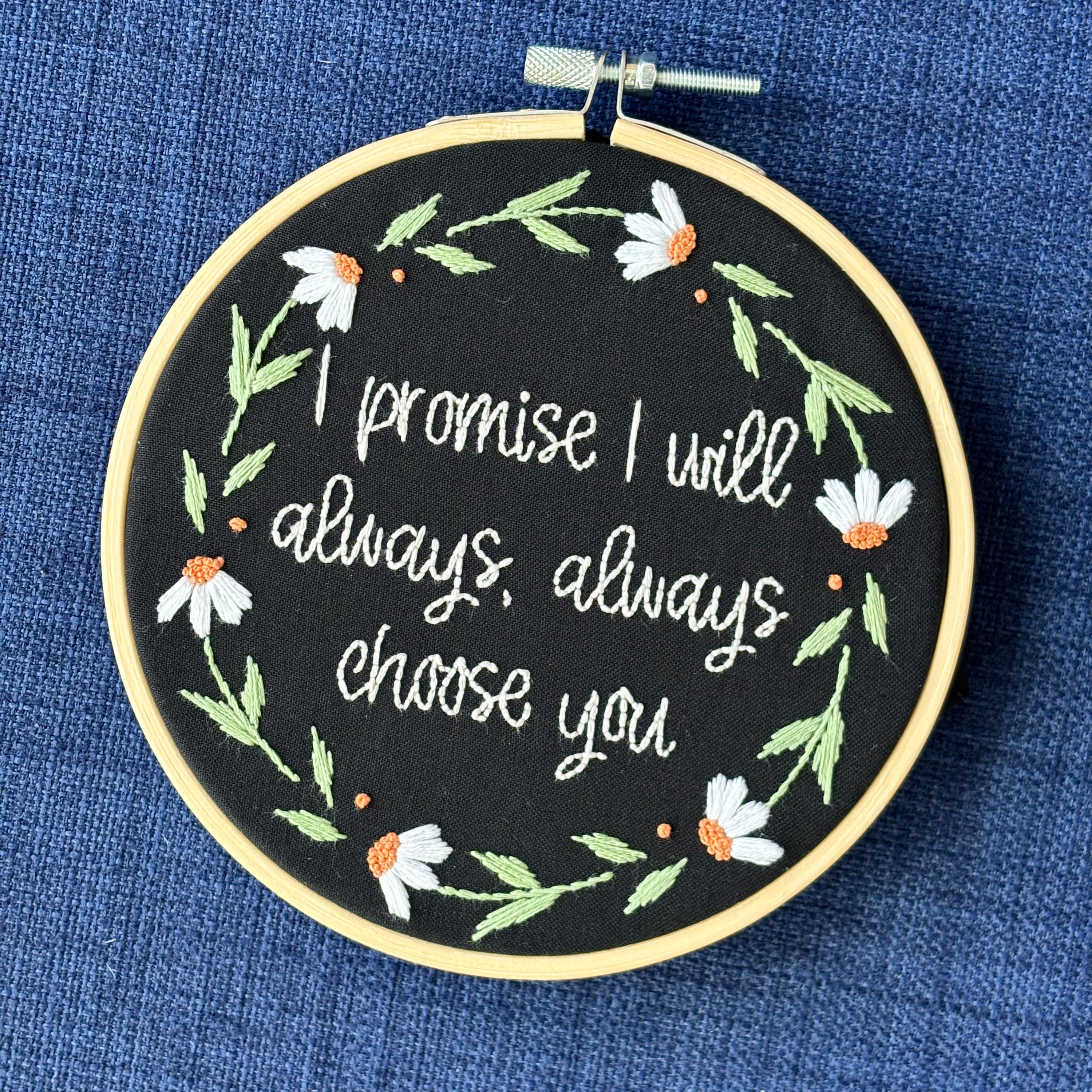 "I promise I will always, always choose you."  Quote hand embroidered in handwriting font in the palest blush pink. Surrounding the text is a floral wreath of white daisies with orange centers. Sewn on black cotton. Framed in a light wood embroidery hoop as wall art.