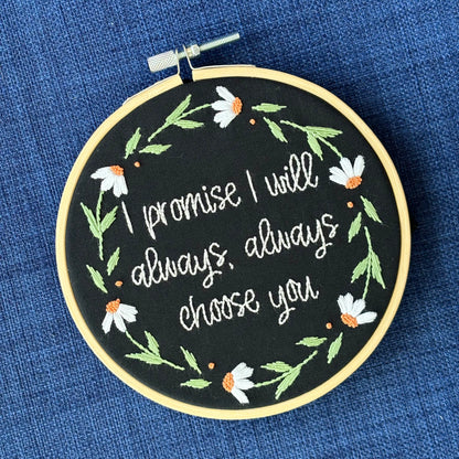 "I promise I will always, always choose you."  Quote hand embroidered in handwriting font in the palest blush pink. Surrounding the text is a floral wreath of white daisies with orange centers. Sewn on black cotton. Framed in a light wood embroidery hoop as wall art.