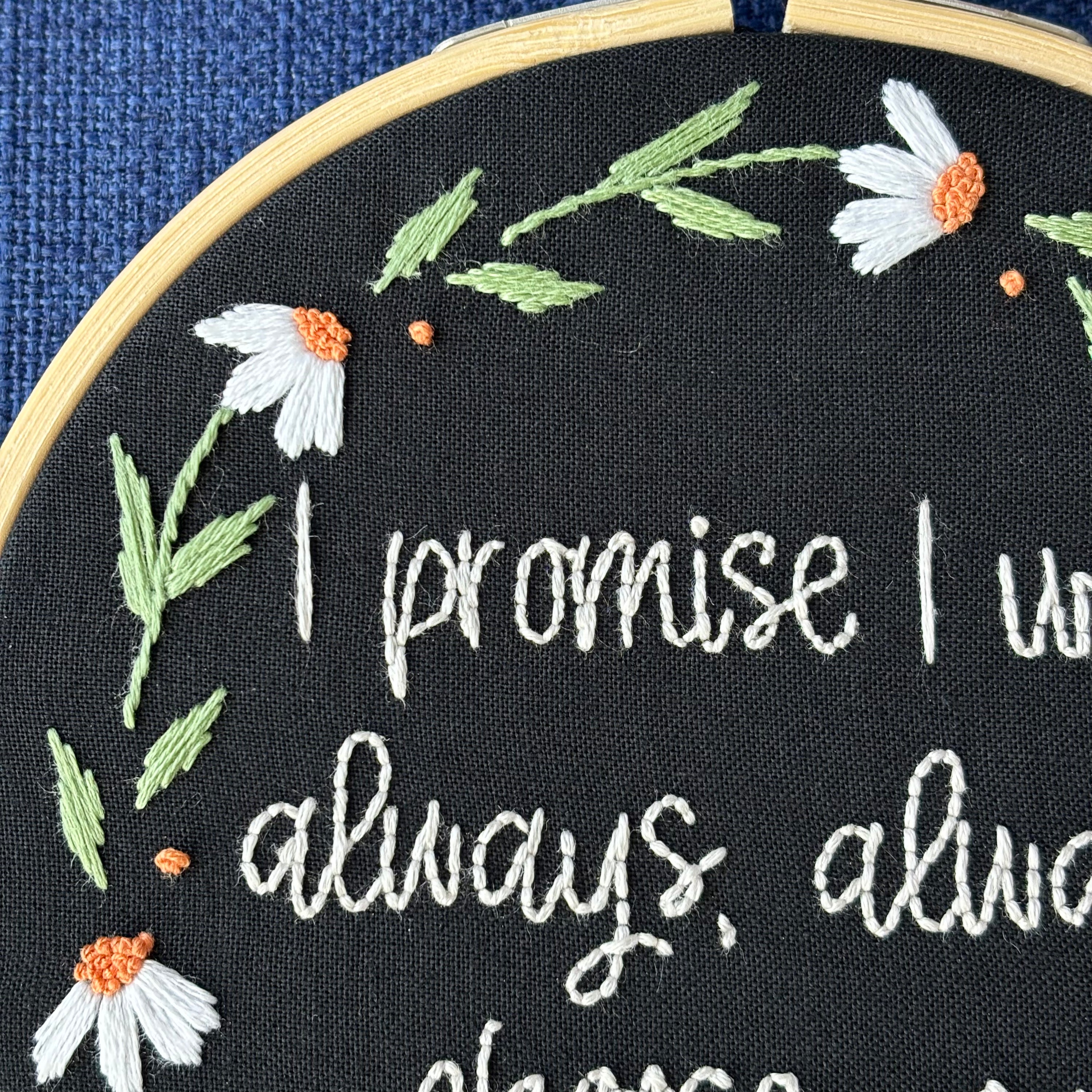 "I promise I will always, always choose you."  Quote hand embroidered in handwriting font in the palest blush pink. Surrounding the text is a floral wreath of white daisies with orange centers. Sewn on black cotton. Framed in a light wood embroidery hoop as wall art.