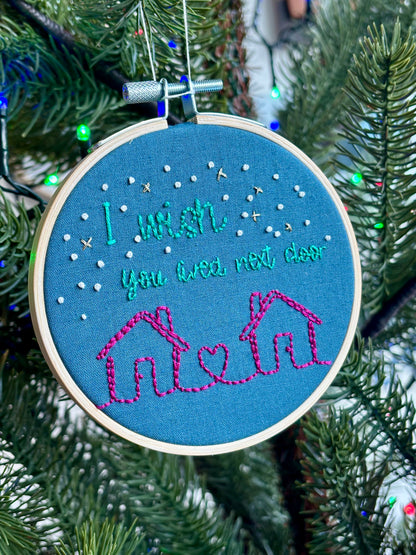 PDF Pattern. Two line drawing houses connected with a heart, hand stitched in bright pink thread. The sky has white and gold stars. Teal text reads, "I wish you lived next door." Cotton fabric is medium blue. Finished Christmas ornament is framed in a light wooden embroidery hoop and hung on a Christmas tree.