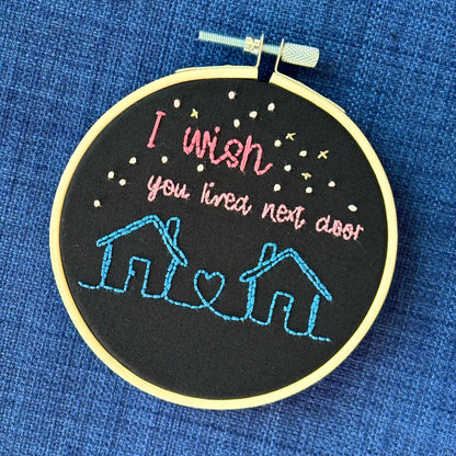 Two line drawing houses connected with a heart, hand stitched in blue thread. The sky has white and gold stars. Pink text reads, "I wish you lived next door." Fabric is black cotton. Finished Christmas ornament is framed in a light wooden embroidery hoop. 