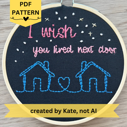PDF Pattern created by Kate, not AI. Two line drawing houses connected with a heart, hand stitched in blue thread. The sky has white and gold stars. Pink text reads, "I wish you lived next door." Fabric is black cotton. Finished Christmas ornament is framed in a light wooden embroidery hoop. 