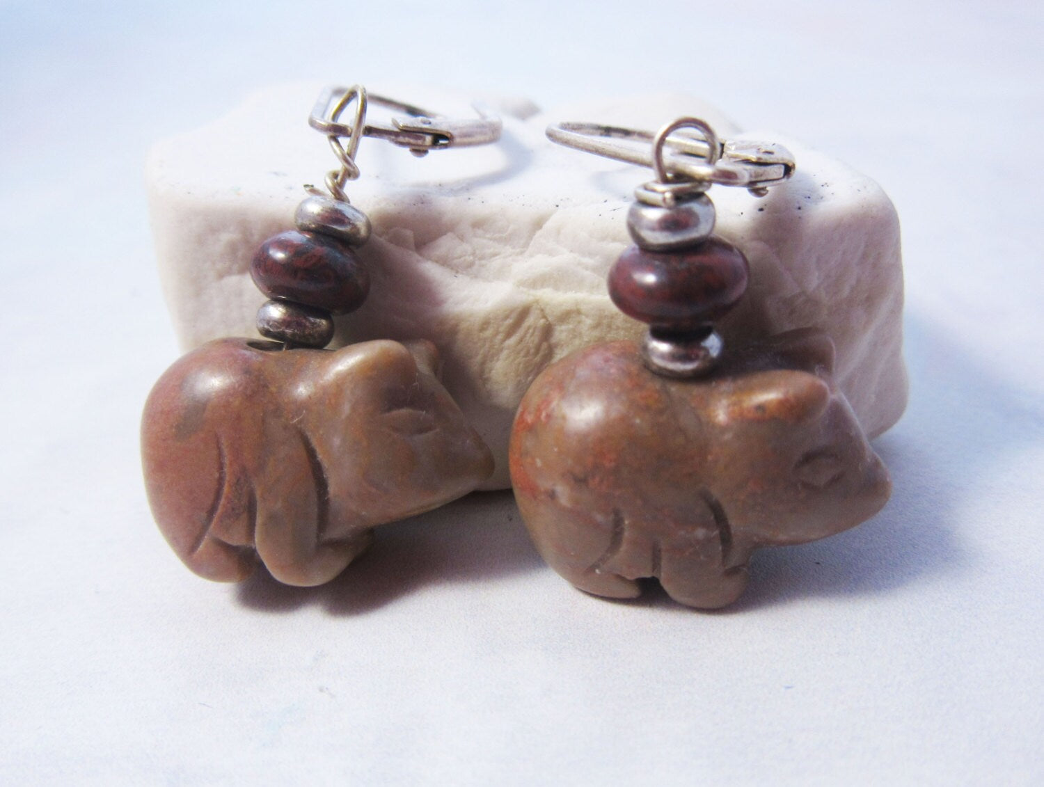 A pair of Carved Stone Pig Earrings featuring small pig-shaped stone pendants on sterling silver leverbacks, accented with round beads and silver spacers, displayed on a white stone background.