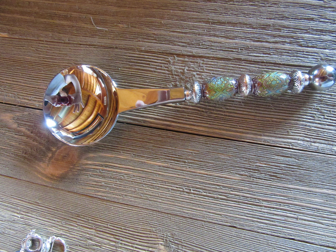 The Artisan Crafted Herb &amp; Bath Salt Spoon Color Changing Mood Bead Scoop, with its vibrant patterns, silver accents, and decorative handle, rests on a wooden surface. Its round bowl shines beautifully, reflecting the surroundings.