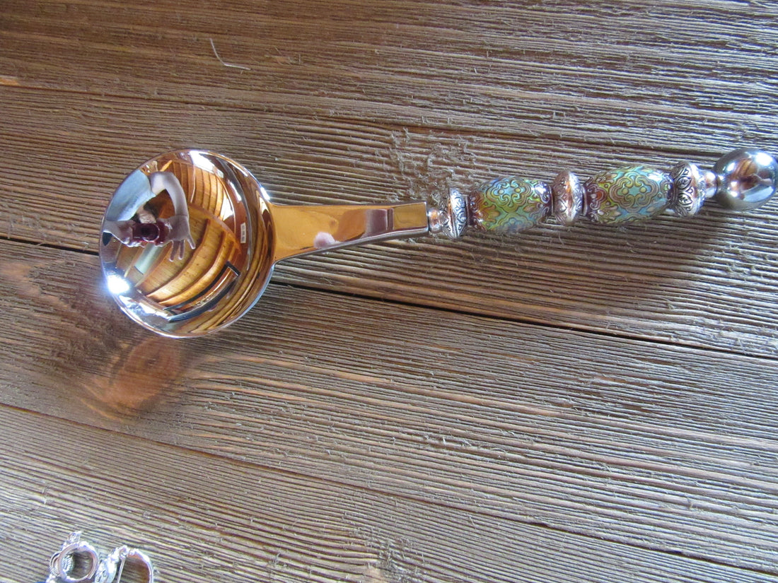 The Artisan Crafted Herb &amp; Bath Salt Spoon Color Changing Mood Bead Scoop, with its vibrant patterns, silver accents, and decorative handle, rests on a wooden surface. Its round bowl shines beautifully, reflecting the surroundings.