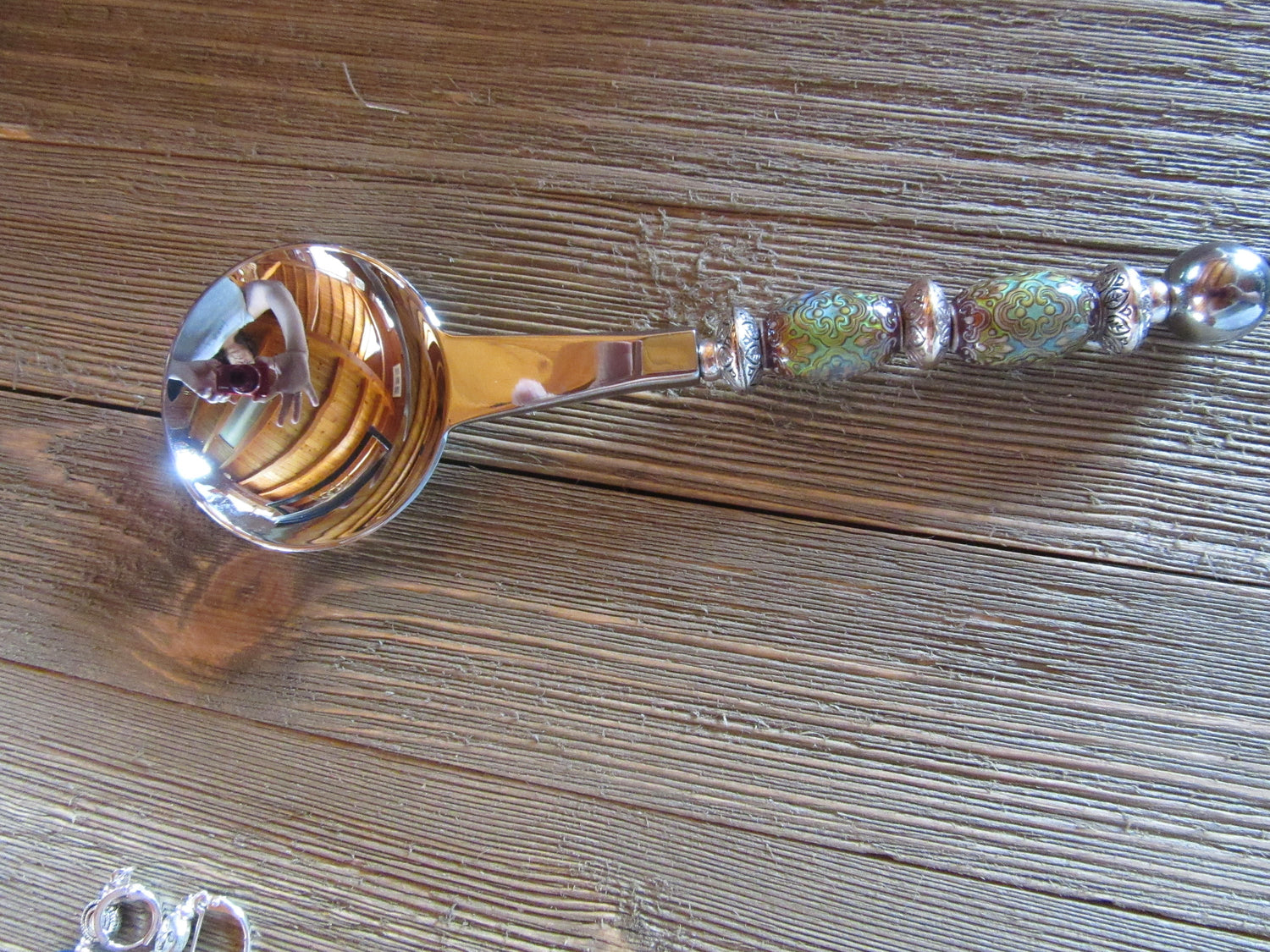 The Artisan Crafted Herb &amp; Bath Salt Spoon Color Changing Mood Bead Scoop, with its vibrant patterns, silver accents, and decorative handle, rests on a wooden surface. Its round bowl shines beautifully, reflecting the surroundings.