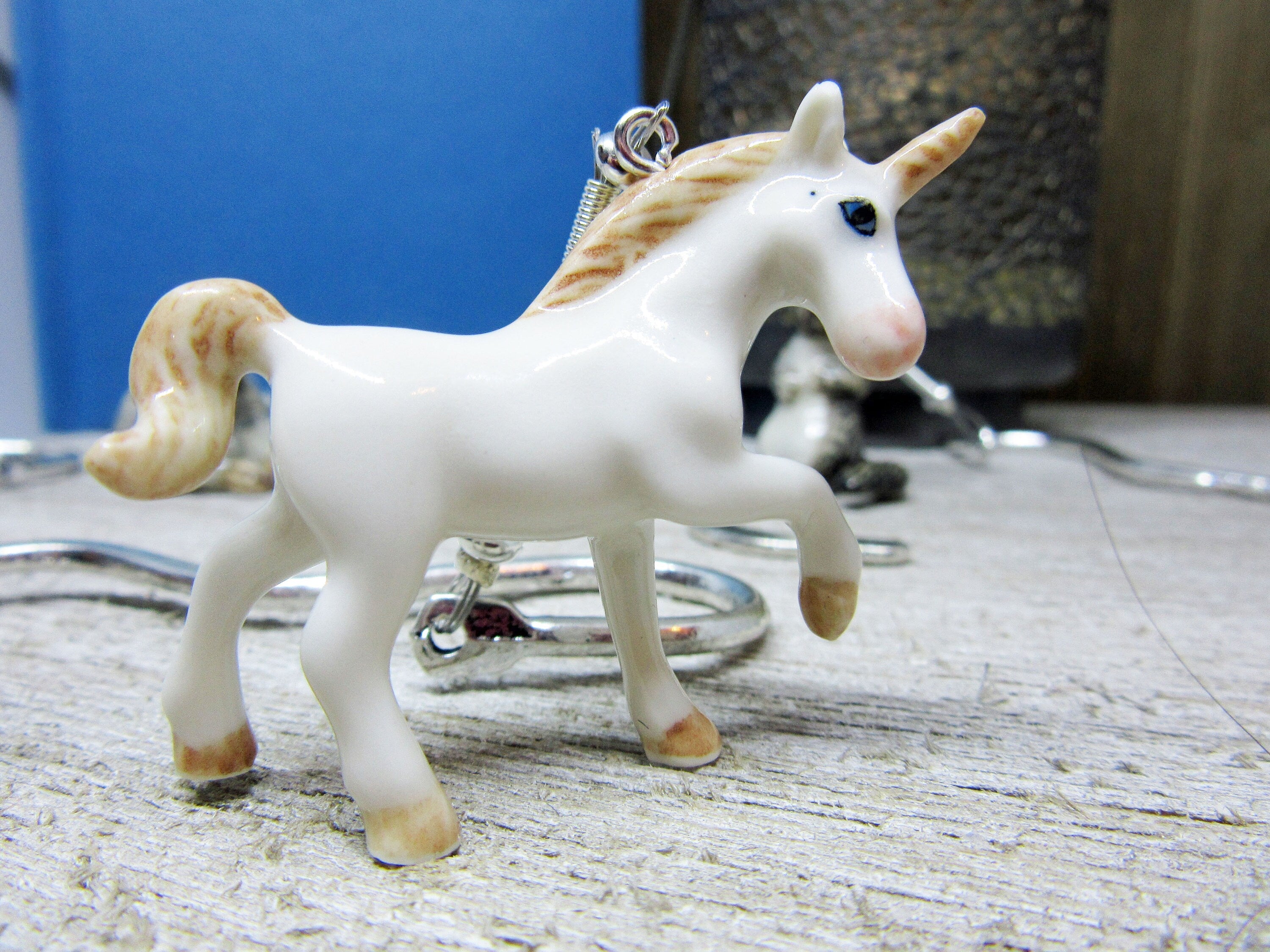 The Porcelain Unicorn Bookmark—an elegant metal bookmark featuring a petite ceramic unicorn with a golden mane and horn—rests on a light wooden surface, accented by silver jewelry and set against a blurred blue-gray background.