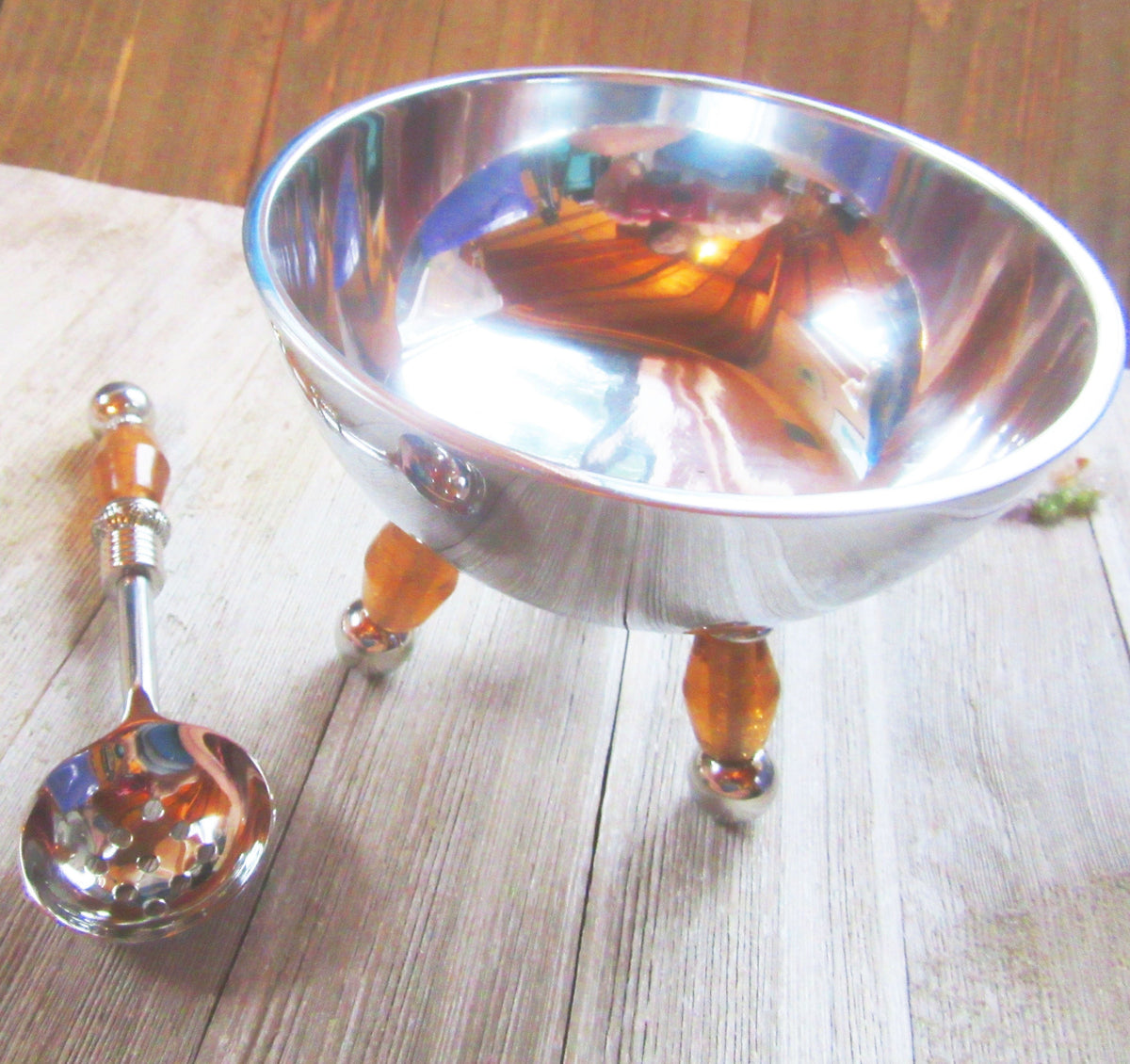 A Bowl set featuring a stainless steel serving bowl with peach art glass legs sits on a wooden surface next to a matching spoon with an orange handle. The bowl reflects light and surroundings. Includes slotted spoon, condiment and dip bowls.