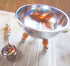 A Bowl set featuring a stainless steel serving bowl with peach art glass legs sits on a wooden surface next to a matching spoon with an orange handle. The bowl reflects light and surroundings. Includes slotted spoon, condiment and dip bowls.