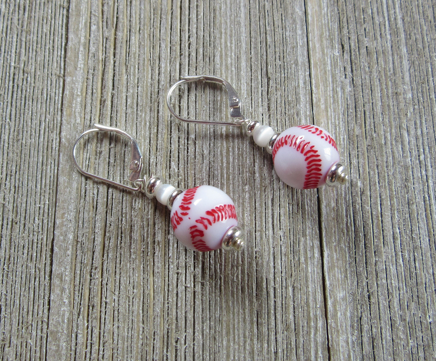Sterling Silver Lever Back Baseball Earrings with red stitching, designed to resemble baseballs, are displayed on a wooden surface.