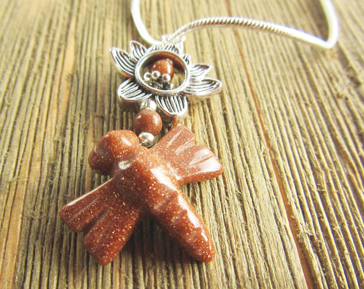 The Goldstone Dragonfly Necklace: Water Lily Pendant hangs from a silver chain and features a sparkling brown design, shown on a textured wooden surface.