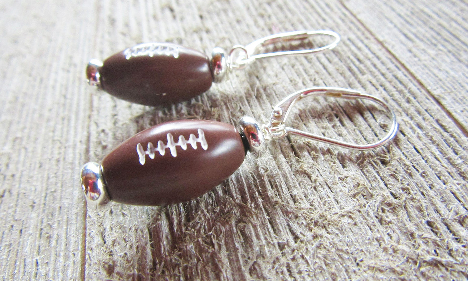 Football Earrings: Sterling Silver Sports Jewelry Gift for the big game, featuring small brown footballs with white stitching on sterling silver hooks, displayed on a textured wooden surface.