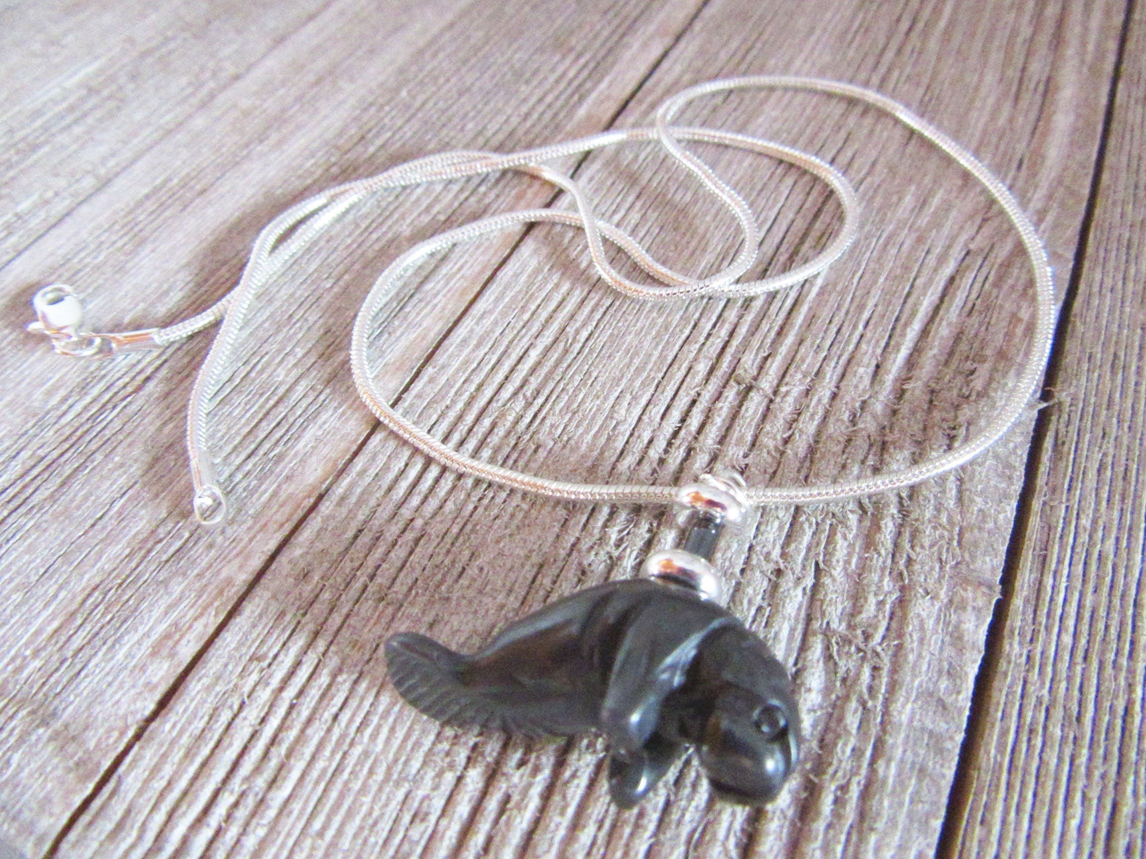 The Handcrafted Manatee Necklace features a dark stone pendant and hematite beads on a silver snake chain, elegantly displayed on rustic wood.