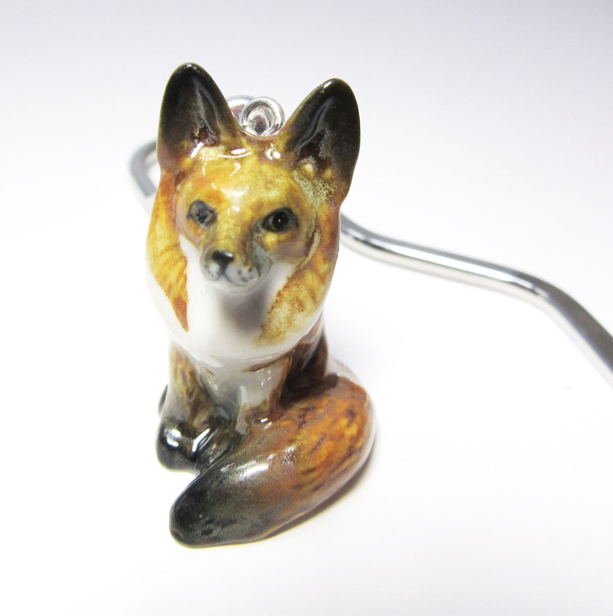 A Handmade Painted Fox Bookmark—a small, glossy ceramic fox with a curled tail—is perched on a white surface, its silver metal hook partly visible. This unique reading accessory makes an ideal gift for book lovers.
